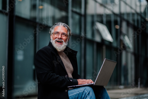 Portrait of a senior businessman using laptop and sitting on a bench outside