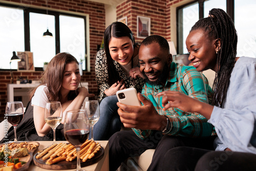 Young african american couple showing photos on cell phone screen to different ethnicities women  everyone smiles. Multicultural group friends sitting  looking mobile  smiling  talking  discussing.