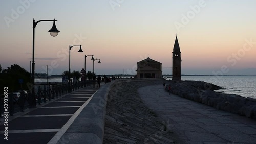 People stroll at dawn on the seafront of Caorle, in the background the sanctuary 