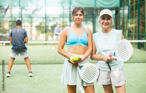 Portrait of friendly young and senior women standing on padel tennis court together during training outdoors. © JackF