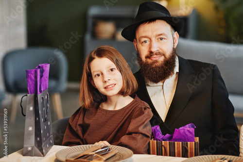 Portrait of modern jewish man with daughter looking at camera while sitting at dinner table in cozy home setting photo