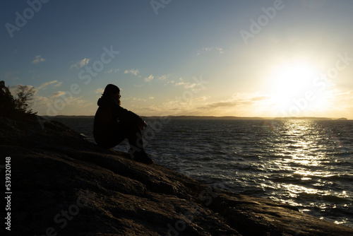 Bearded Man with a beanie hat sitting on a cliff by the seaside looking towards the sunset