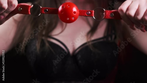 Young girl in black lingerie close-up holds a red bdsm gag on a black background. Pulls on the gag, brings it to his mouth. BDSM is an outfit for adult sex games
 photo