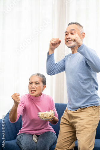 Excited mature couple, Senior man and woman watching tv, senior sport fans celebrating favorite team victory, sitting on cozy couch and eating popcorn snack at home, enjoying weekend. Happy senior.