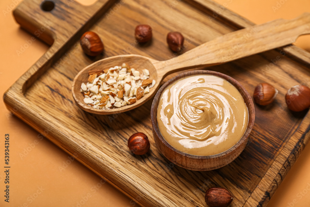 Wooden board with bowl of tasty nut butter and hazelnuts on color background, closeup Wooden board with bowl of tasty nut butter and hazelnuts on color background, closeup