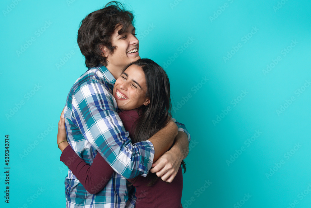Happy couple in love hugging, embracing each other; casual wear, teenage years, fresh relationship; teal studio background Happy couple in love hugging, embracing each other; casual wear, teenage years, fresh relationship; teal studio background