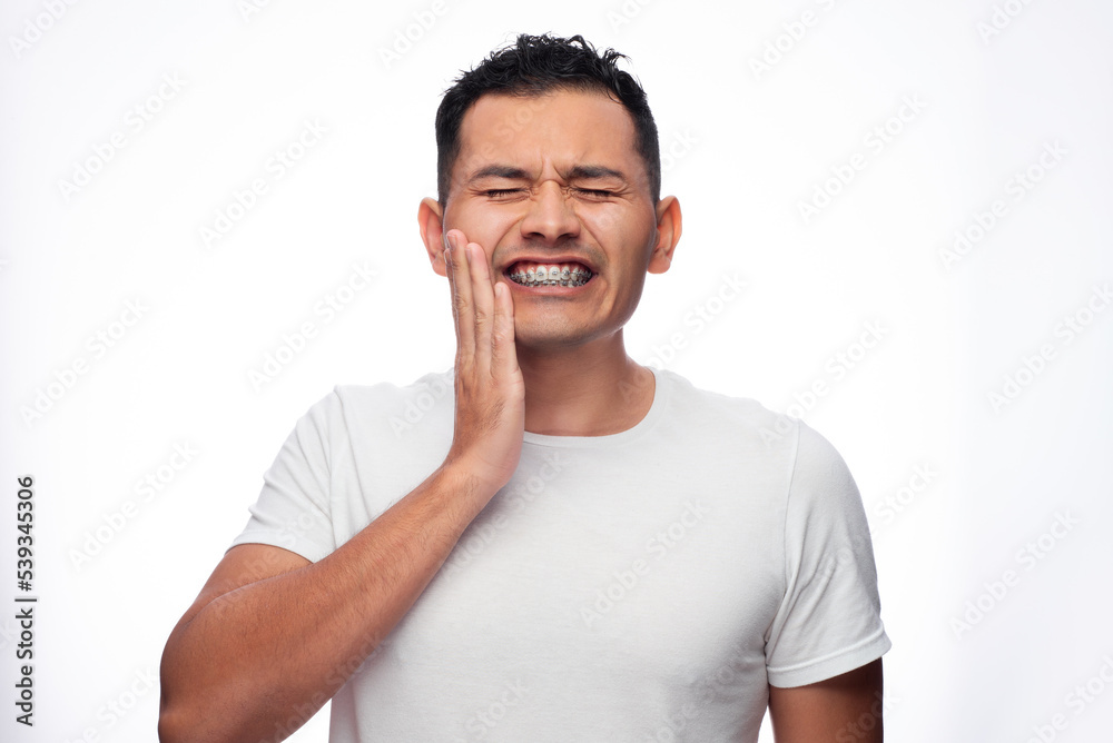 Hispanic man in white shirt and braces grimaces in pain on his face and holds his face by his orthodontic treatment brackets Hispanic man in white shirt and braces grimaces in pain on his face and holds his face by his orthodontic treatment brackets
