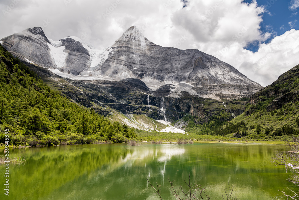 Horizontal image of the beautiful Pearl Lake or Zhuoma La Lake and snow mountain in summer in Yading Nature reserve, Sichuan, China. Beautiful nature landscape. Shades of green, blue sky with clouds