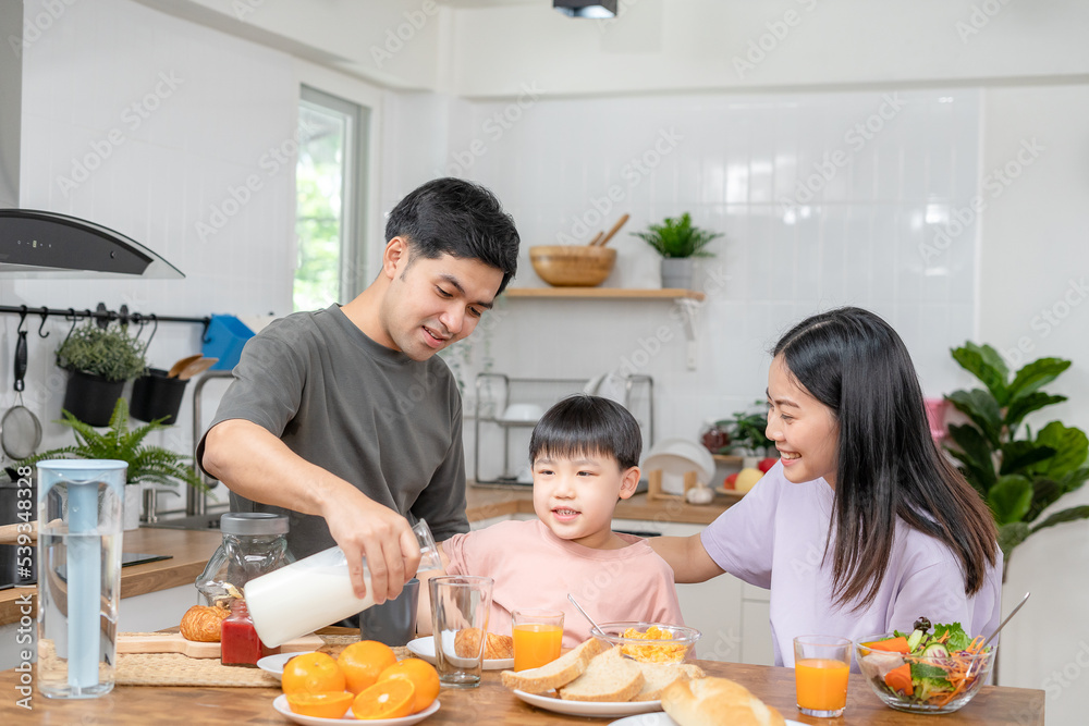 Asian family breakfast at home. Parents and children enjoy eating together, talking with laughter and good atmosphere. Father plays with son playfully at kitchen table.