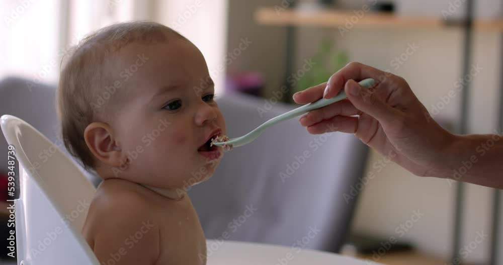 Baby girl 8 months old in high chair being spoon fed by mother - side profile Baby girl 8 months old in high chair being spoon fed by mother - side profile