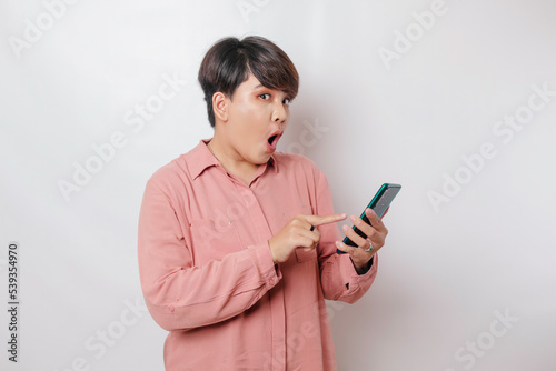 Surprised short-haired Asian woman wearing a pink shirt pointing at her smartphone, isolated by a white background