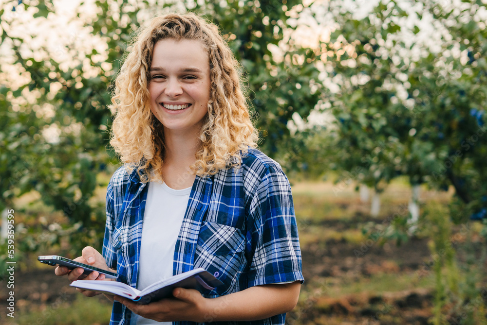 Woman specialist writing on notepad while using smart phone, working in plum orchards. Online business network technology. Agricultural business concept Woman specialist writing on notepad while using smart phone, working in plum orchards. Online business network technology. Agricultural business concept