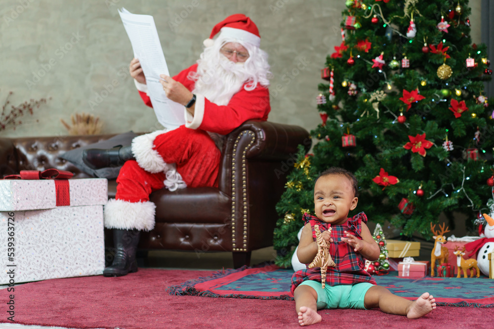 Senior man in Santa Claus costume reading newspaper on sofa watching African American baby girl playing and crying on the floor. Christmas holiday season