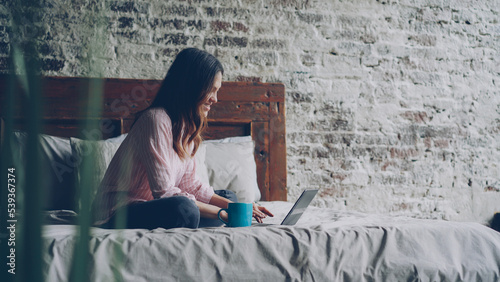 Attractive young woman in casual clothes is typing on laptop and smiling while sitting on bed in nice loft style bedroom at home. Modern technology and millennials concept.
