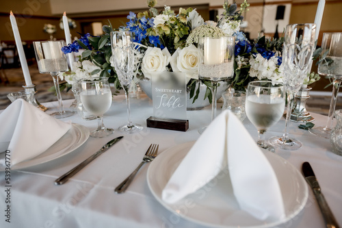 folded napkins on sweetheart table in ballroom with wedding floral centerpiece on white table cloth surrounded by place settings and glassware. blue hydrangeas white roses horizontal no people
