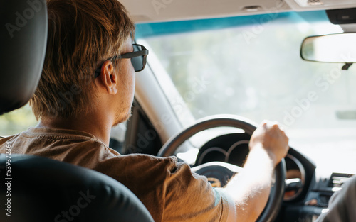 Caucasian man in sunglasses driving a car and looking ahead at road, close-up. Back view of driver holding steering wheel, selective focus