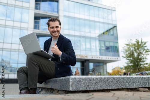 a male freelancer with a laptop on the background of a modern office building looks at the camera in a wide smile