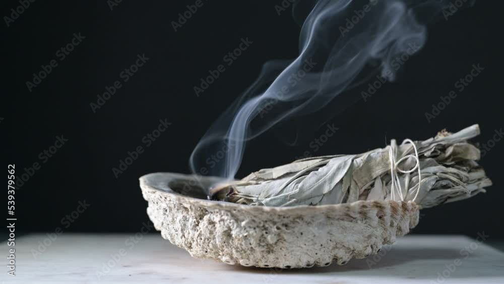 Burning sage in a shell on a wood table with black background showing sage smoke