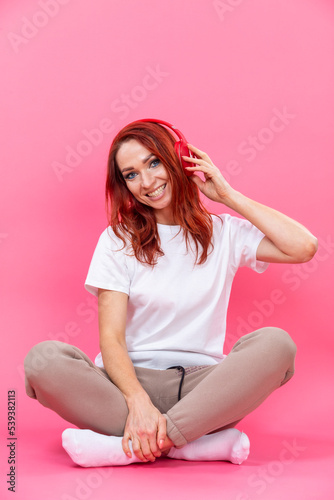 Winsome cheerful stylish young woman wearing white shirt sitting on hunkers isolated over pink background while listening to music with wireless headphones