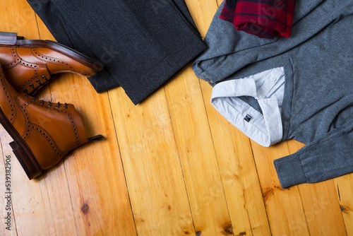 Clothing Concepts. View Flat Lay of Tan Brogues Boots With Jumper, White Shirt, Sweater, Pair of Herringbone Trousers on Wooden Surface Background.