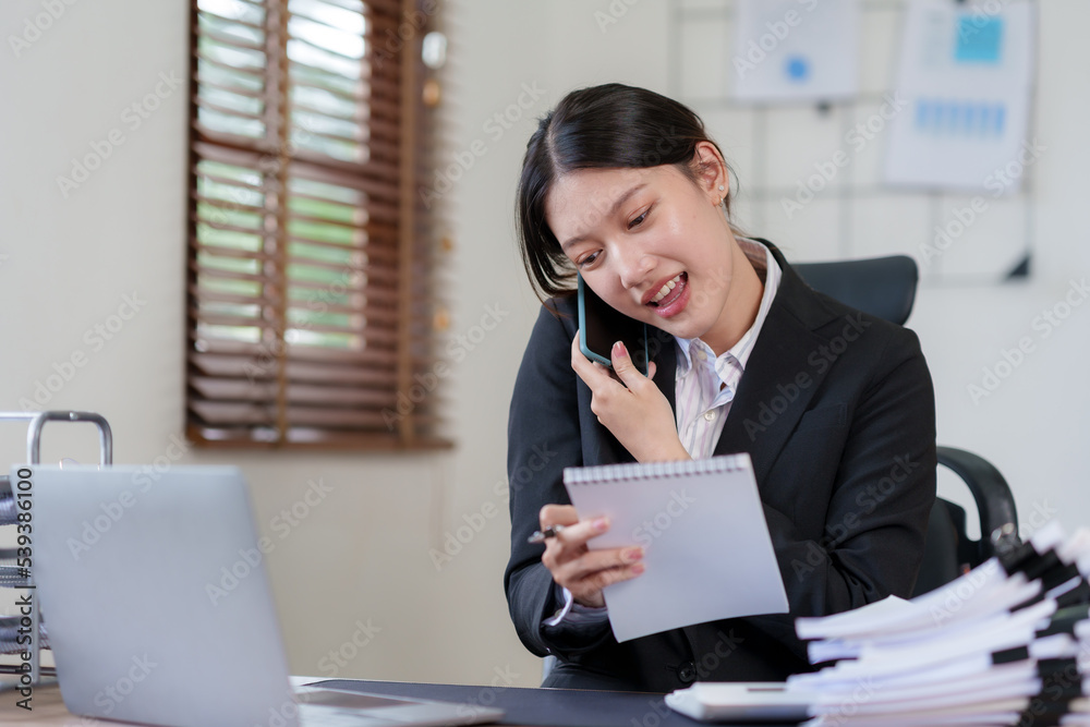 Happy Asian businesswoman smiling and talking on the phone with a client in the office with a bunch of papers on the desk. Happy Asian businesswoman smiling and talking on the phone with a client in the office with a bunch of papers on the desk.