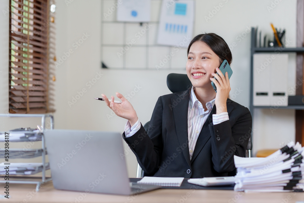 Happy Asian businesswoman smiling and talking on the phone with a client in the office with a bunch of papers on the desk. Happy Asian businesswoman smiling and talking on the phone with a client in the office with a bunch of papers on the desk.