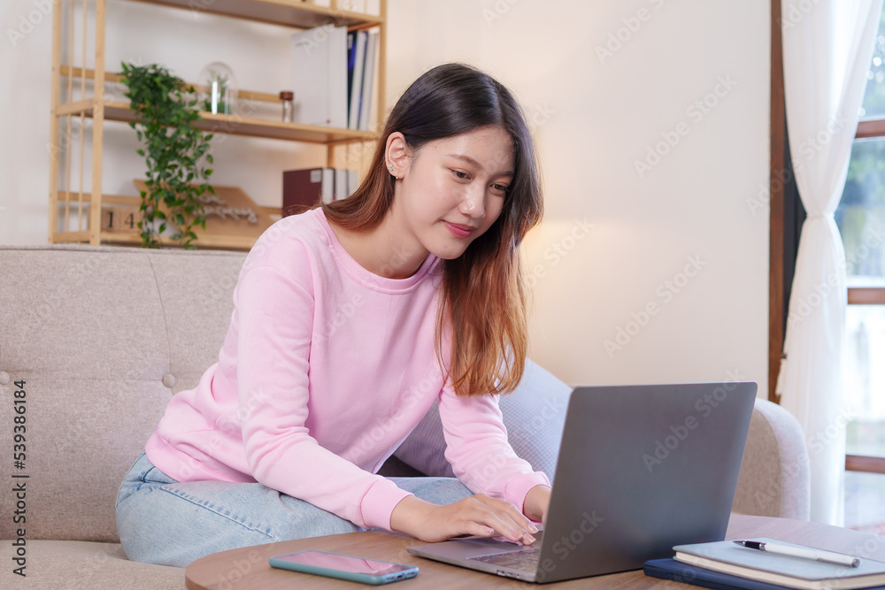 Bright, cute and happy Asian female student study online in the living room using laptop computer in a relaxed manner. Young Asian woman using laptop at home.