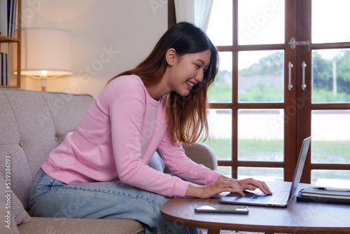 Bright, cute and happy Asian female student study online in the living room using laptop computer in a relaxed manner. Young Asian woman using laptop at home.