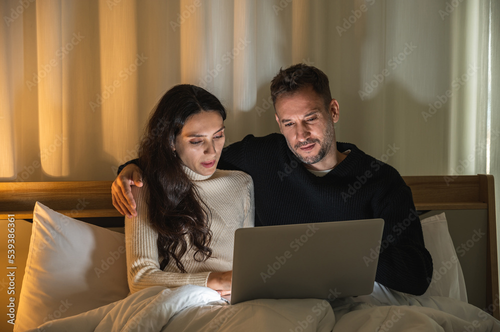 Caucasian couple siting together on bed in dark room looking at a laptop screen using computer to search for something.