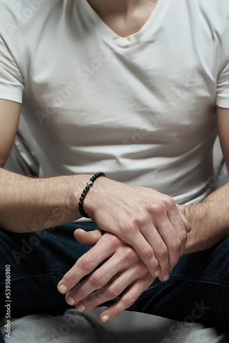 Cropped close-up shot of man s hands with a black stone bracelet with a miniature hematite cross. The man is sitting and holding his hands with stone bracelet together. Front view.