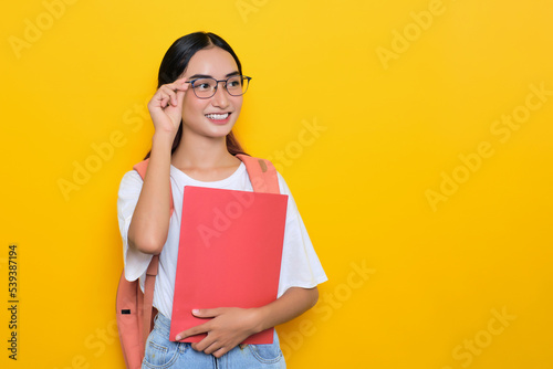 Cheerful pretty young student girl wearing backpack and glasses holding books isolated on yellow background