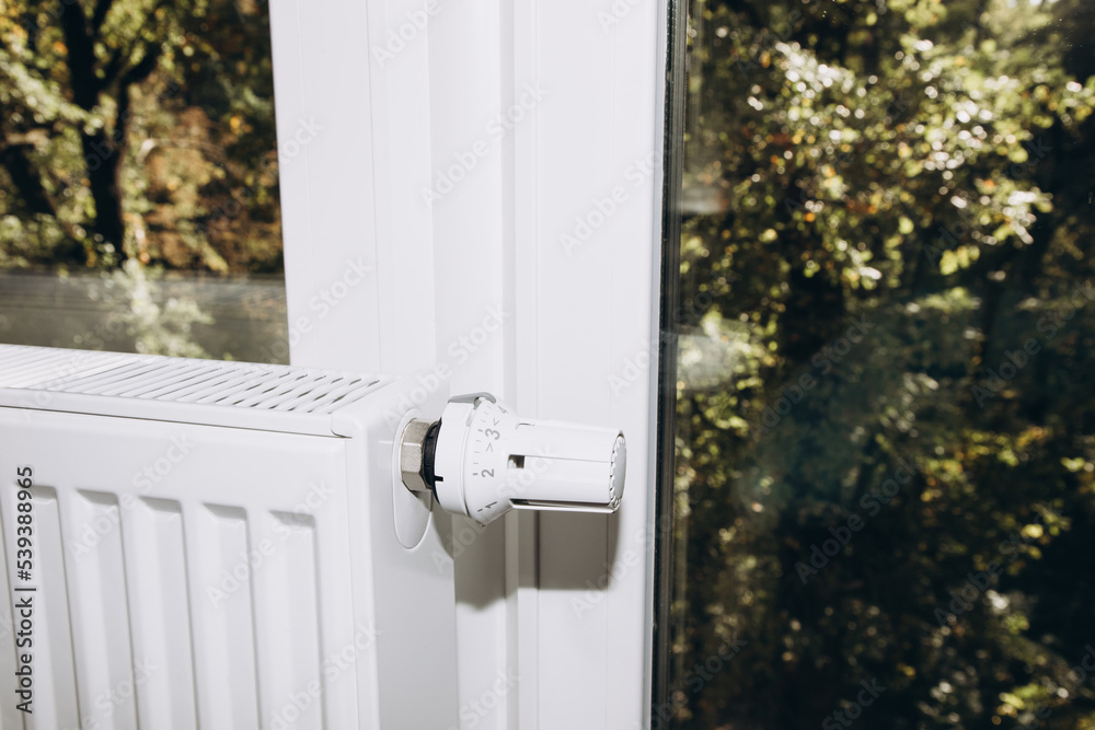 White metal battery at home near a panoramic window with a thermostat close-up. Apartment heating White metal battery at home near a panoramic window with a thermostat close-up. Apartment heating
