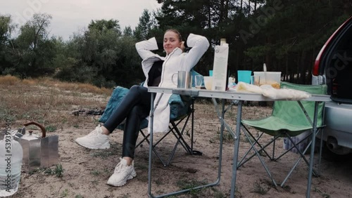 camping, a beautiful woman is sitting at a tourist table in an armchair and enjoying unity with nature in a pine forest.