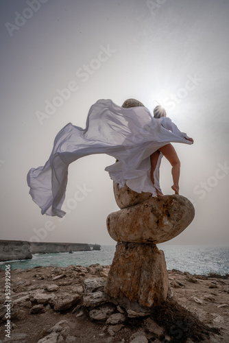 A woman stands on a stone sculpture made of large stones. She is dressed in a white long dress, against the backdrop of the sea and sky. The dress develops in the wind.