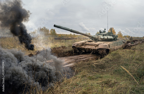 Heavy Military Tank on the battlefield at a hot spot. A tank moves through a field amidst explosions. Dramatic view on the tank moving in a cloud of smoke and dust.