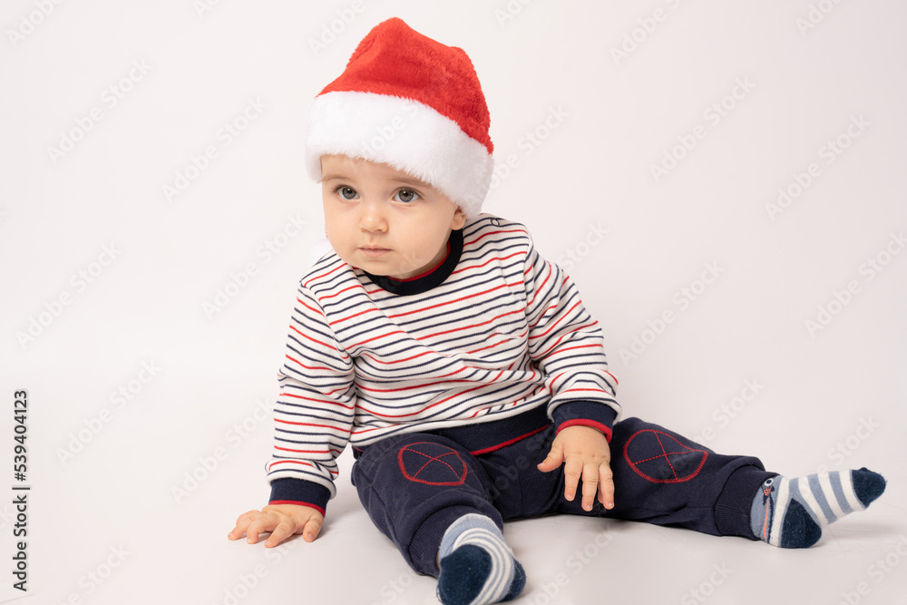 Adorable child is sitting on floor, wearing red Christmas cap, isolated over white background. Adorable child is sitting on floor, wearing red Christmas cap, isolated over white background.