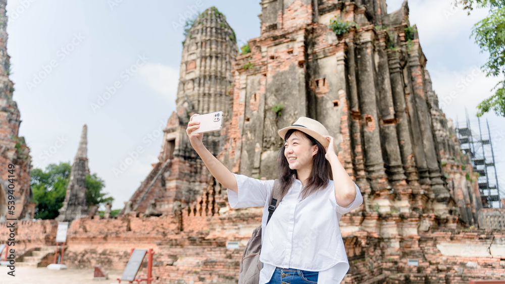 Young Asian woman traveler using mobile phone taking selfie while solo travel on ayuthaya temple thailand in summer sunny day. Cheerful female enjoy outdoor lifestyle in holiday