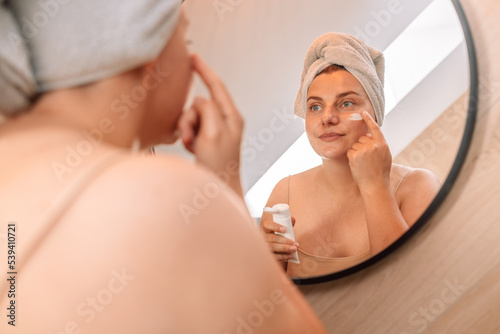 Cute beautiful young girl sits after a shower with a white towel on her head and smears her face with cream.