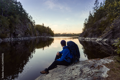 A caucasian man with a backpack sitting on a rock in a forest next to a lake at sunrise.