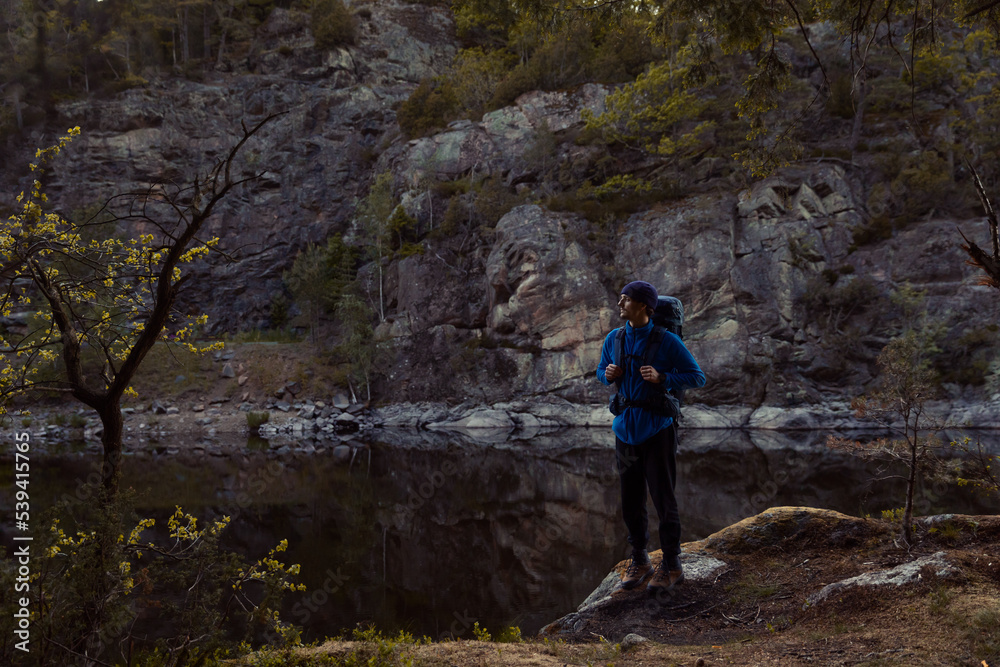 A man with a backpack standing in a forest on a rock next to a lake at sunrise.