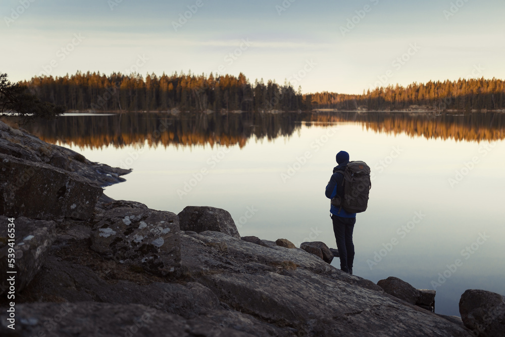 A caucasian man with a backpack standing on a rock in a forest next to a lake at sunrise.