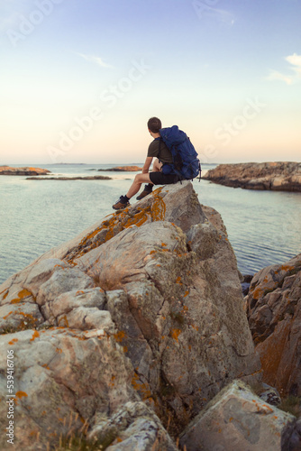 A caucasian man sitting on a rock by the sea with backpack overlooking the sea at sunrise.