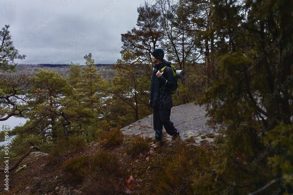 A caucasian man with a backpack standing on a rock in a forest on a rainy day watching over a lake.