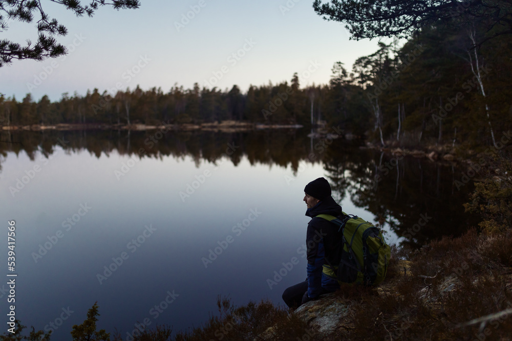 A caucasian man sitting on a rock with a backpack by a lake in a forest at sunset.