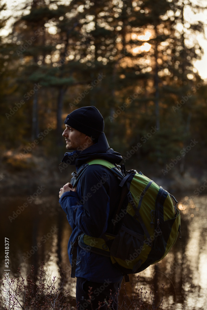 A caucasian man standing by a lake in a forest with a backpack at sunset.