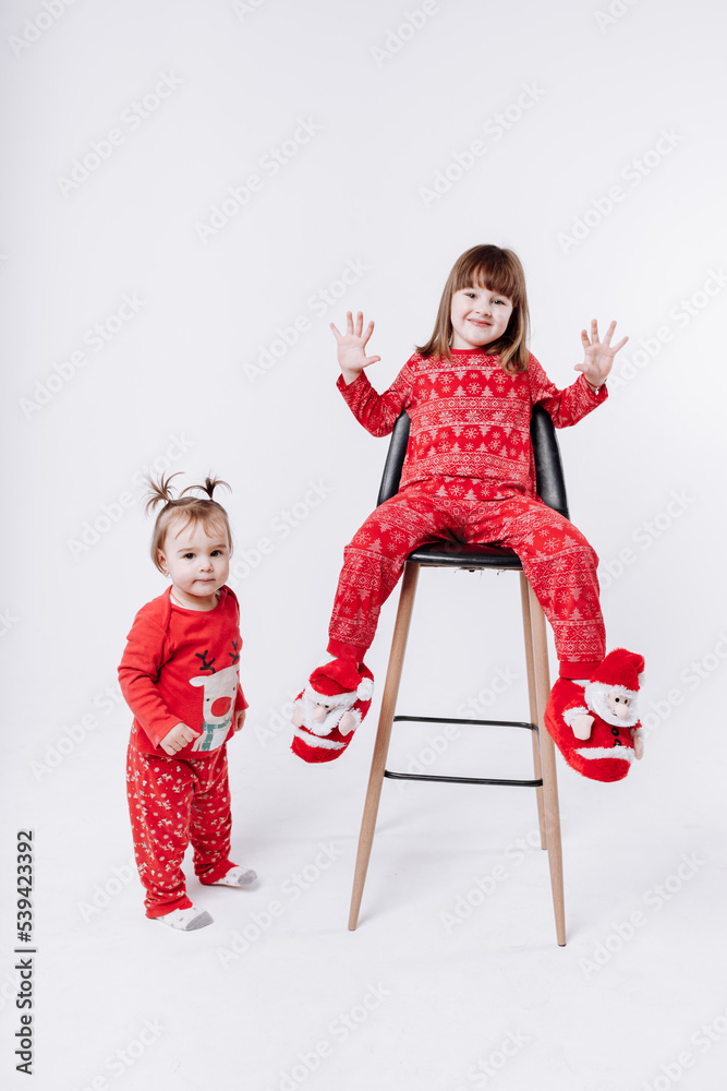 Merry Christmas and Happy family Holidays. Cheerful cute children girls wearing in Xmas red pajames having fun on white background. Smiling older sister sitting on chair and playing. Loving sibling