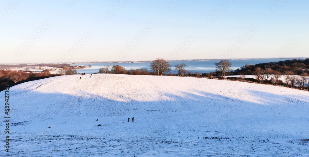 Danish winter landscape with white fields and hills close to the sea and children sledging in the snow