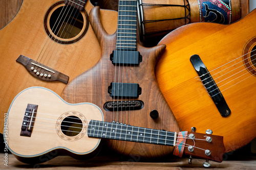 Acoustic and electric guitars, ukulele and djembe standing side by side on a wooden background.