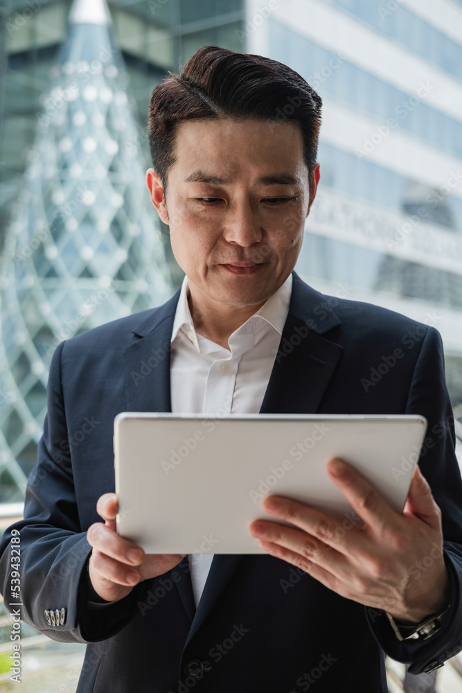 Smiling Businessman Using A Digital Tablet While Standing At City Street With Modern Office Buildings In The Background Smiling Businessman Using A Digital Tablet While Standing At City Street With Modern Office Buildings In The Background