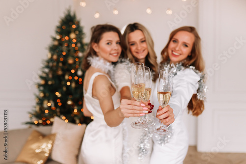 Portrait of happy three female friends in fashionable shiny dresses holding glasses of champagne laughing and celebrating the Christmas holiday in a decorated house. Selective focus